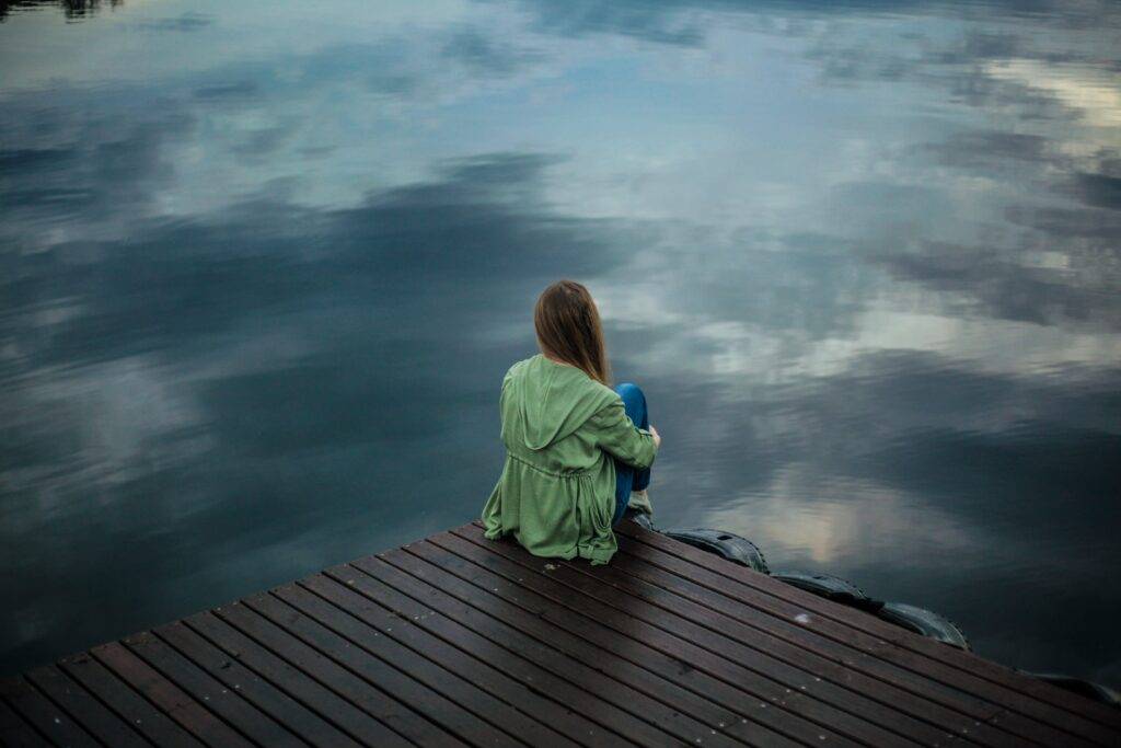 Vulnerability encompassing a girl while she gazes out on the lakeside docks on a cloudy day.